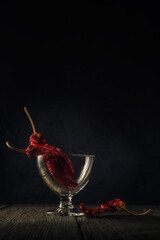 dried red hot chili peppers in a glass goblet and on a wooden table against the background of a black concrete wall. side view. dark artistic minimalist still life with copy space