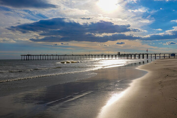 Long pier seen extending out into a calm ocean from a deserted beach with a dramatic cloudscape just before sunset
