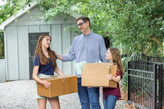 A Father And His Two Daughters Holding Moving Boxes Outside On A Gravel Driveway Near A Storage Building