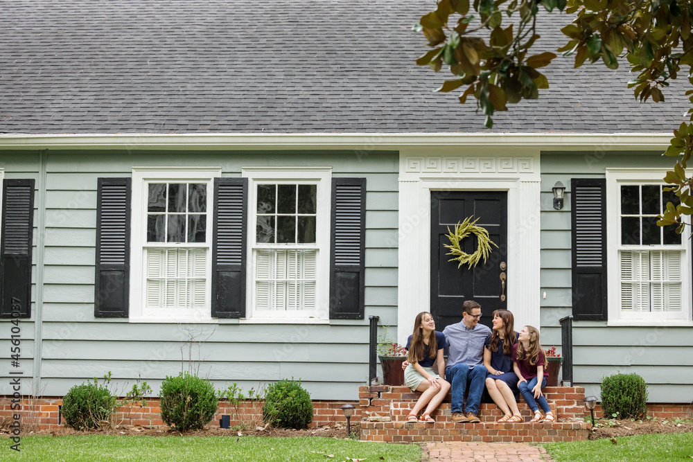 Wall mural A family with a mother, father, and two daughters sitting outside on the brick steps of a front porch of a small blue cottage house