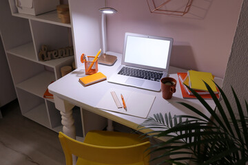 Stylish workplace with laptop, stationery supplies, cup and glowing lamp near light wall