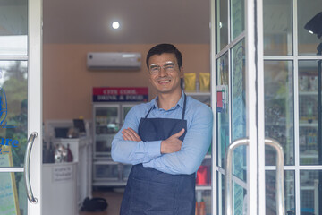 Young male owner expresses his warm welcome with his hand to customer for his local startup bakery or coffee shop in front of the shop. Smiling attractive owner with glasses stands for welcoming.