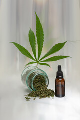 A bottle of cannabis oil next to a bottle of watered dry herb in front of a marijuana leaf on gray glossy background