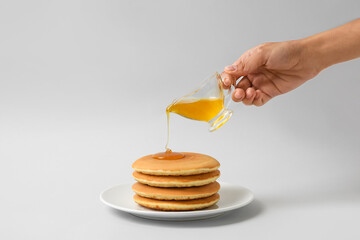 Woman pouring honey onto plate with tasty pancakes on light background