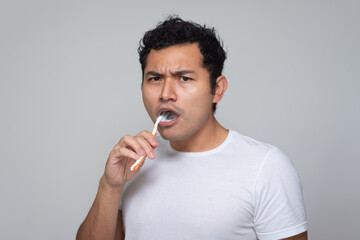 Mexican man with Latino Hispanic appearance brushes his teeth with an orange toothbrush, young man with Chinese hair in white shirt with gray background back