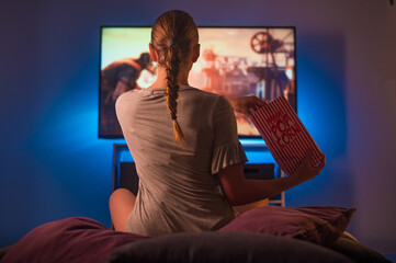 A blonde girl watches TV with a pack of popcorn in her hand. Cozy home environment. Blue neon...