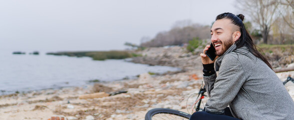 Obraz premium young hispanic latino man smiling on a smart phone call on a beach, with bicycle and sportswear. panoramic photo. copy space