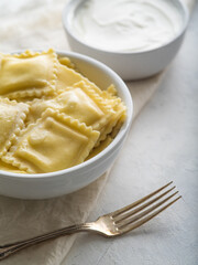 A bowl of ravioli drizzled with butter, a fork and sour cream in the background. Tasty dinner. White tablecloth. High angle view. There is no one in the photo. Home cooking, restaurant, hotel.