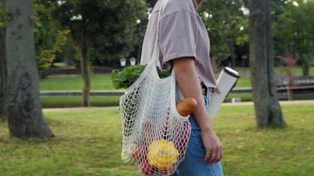 Woman in round glasses walking and carrying white mesh string knitted shopping bag with groceries. Eco friendly, reusable shopping bag. Zero waste and plastic free concept.