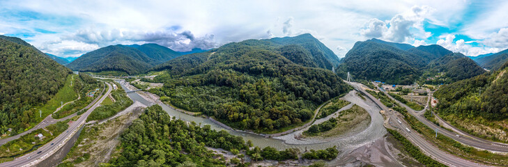 Fototapeta na wymiar a large aerial panorama of the valley of the mountain river Mzymta, surrounded by highways and forested green mountains of the Caucasus. Cable-stayed bridge over the river entering the tunnel