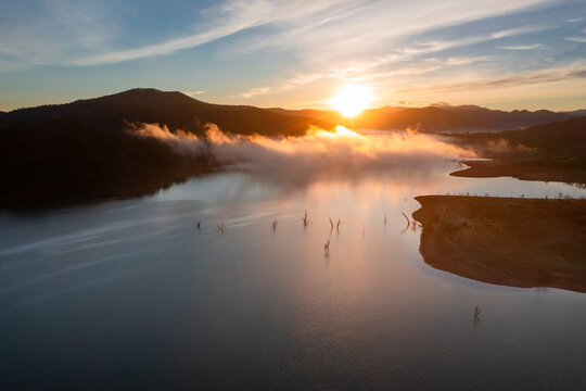 Aerial view of sunrise over mountains surrounding a lake with fog patches