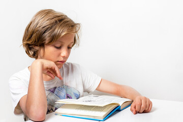 serious boy reading an interesting book at a desk against a background of a white wall, back to school, Authenticity