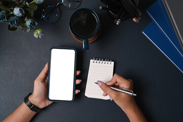 Overhead shot woman holding mobile phone and writing on notepad.