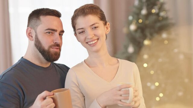 Medium close-up of happy young Caucasian hetero couple standing in foreground of blurred Christmas tree in room lit with warm light, holding mugs with hot chocolate, talking and smiling