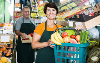 Positive female seller showing basket with vegetables and fruits in supermarket