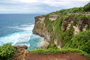 A steep cape with green vegetation and ocean. Rocks crash into the blue waters of the sea near the temple of Pura Luhur Uluwatu in Bali, Indonesia