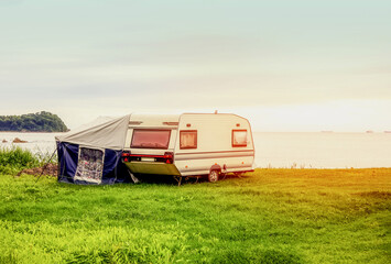 Fototapeta na wymiar Trailer motor home and a tent on the grassy part of the beach at sunset. Leisure mobile camping home for tourists overlooking the blue sea and cape. Adventure relaxing travel on caravan van