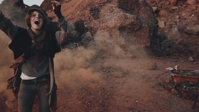 Young happy woman in worn clothes and knitted hat holding colored smoke bomb and dancing with excitement in dystopian world