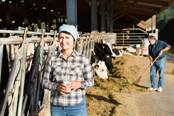 Positive friendly female farmer is standing with glass of cow milk at the farm.