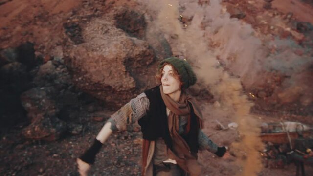 Young excited woman in shabby clothes and knitted hat dancing with colored smoke bomb in dystopian world