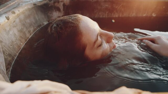 Young woman lying under dirty water in old rusty bath and then getting up at sunset in dystopian world