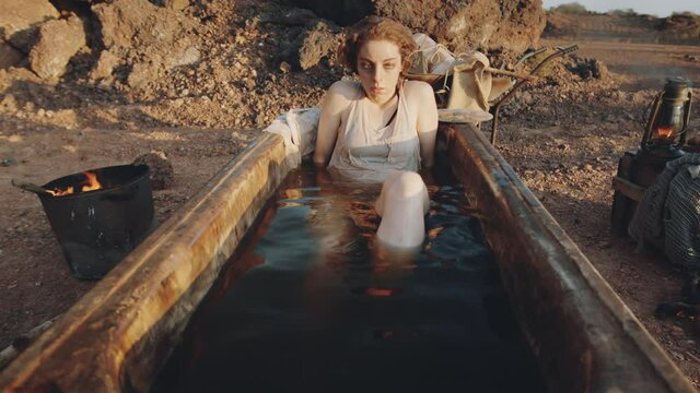 Arc shot of young woman lying in old rusty bath filled with dirty water at campsite in post apocalyptic world