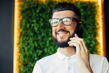 young man in a white shirt and glasses talking on a mobile phone and smiling while sitting in a cafe, Young man sits in a cafe on a coffee break