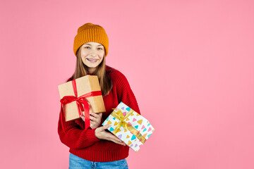 Young smiling happy hipster girl with baces holding present boxes celebrating new year, wearing red knitted sweater and yellow hat. Christmas gifts and winter fashion trend.