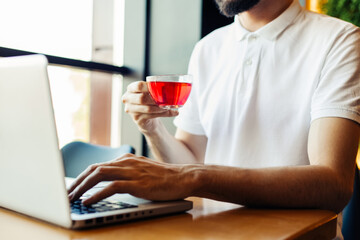 man using a laptop in a cafe and drinking tea while sitting at a wooden table, Male hands are typing on the laptop keyboard