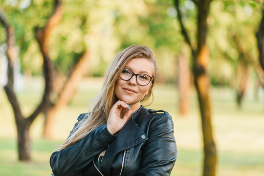 Young blonde woman in glasses for vision posing in the park in sunny weather on the body of a black jacket brutal image