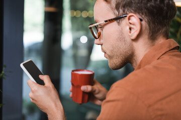 young man in glasses holding cup of coffee and texting on smartphone in cafe
