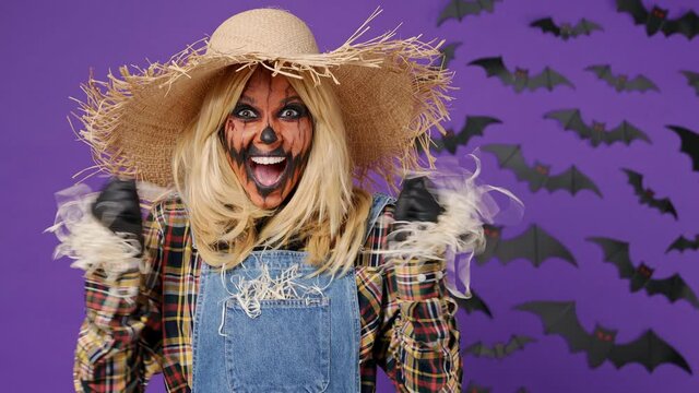 Scary bright vivid young woman with Halloween makeup mask wears straw hat scarecrow costume look camera surprised celebrating clenching fists isolated on plain dark purple background studio portrait