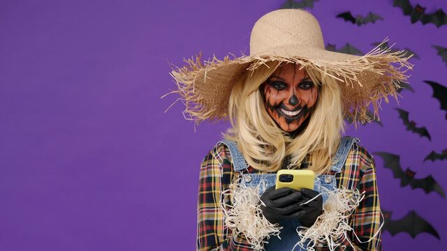 Promoter young woman Halloween makeup mask wears straw hat scarecrow costume hold use mobile cell phone look aside on workspace copy space area isolated on plain dark purple background studio portrait