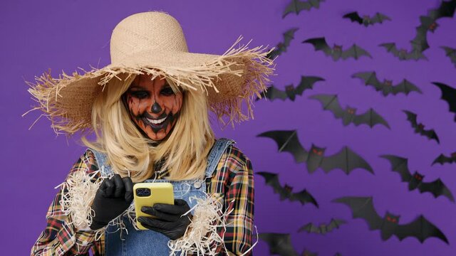 Satisfied smiling young woman with Halloween makeup mask wear straw hat scarecrow costume hold in hand use point on mobile cell phone good news isolated on plain dark purple background studio portrait