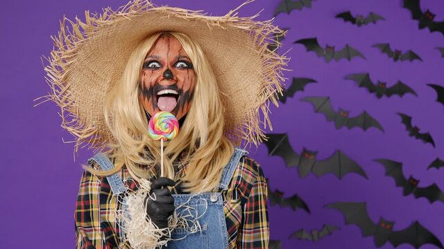 Scary fun young woman with Halloween makeup mask wears straw hat scarecrow costume take candy lick eat taste isolated on plain dark purple background studio portrait. Celebration holiday party concept