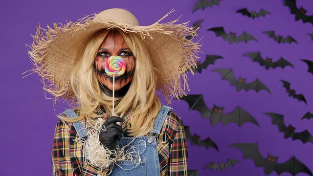 Scary bright smiling happy young woman with Halloween makeup mask wears straw hat scarecrow costume cover face hide behind candy look around isolated on plain dark purple background studio portrait