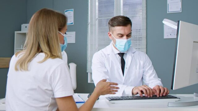 Smiling male professional doctor plastic surgeon wearing protective mask chatting with female patient and agree on surgery. Physician in white uniform and woman patient at medic checkup visit concept