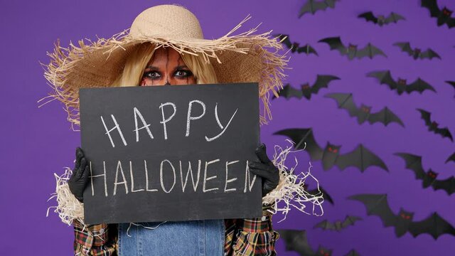 Scary bright vivid young woman with party makeup mask wears straw hat scarecrow costume hold in hand hide behind black poster happy Halloween isolated on plain dark purple background studio portrait