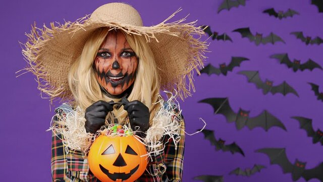 Satisfied excited fun smiling young woman with Halloween makeup mask wear straw hat scarecrow costume raise pumpkin basket with candies licking isolated on plain dark purple background studio portrait