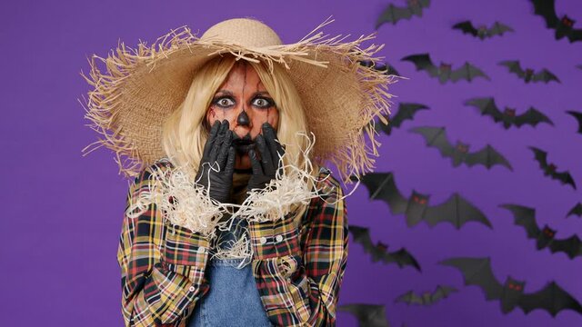 Scared young woman with Halloween makeup mask wear straw hat scarecrow costume look camera covering hiding face with hands peep through fingers isolated on plain dark purple background studio portrait