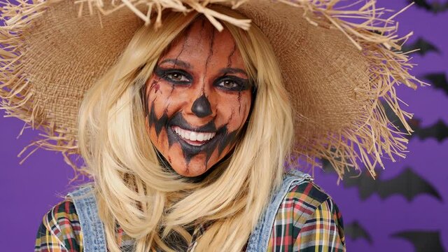 Close up vivid fun young woman with Halloween makeup mask wears scarecrow costume touches straw hat smiling isolated on plain dark purple background studio portrait. Celebration holiday party concept