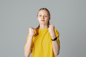 Teen girl with negative facial expression raising fists furiously looking at the camera, isolated on light grey background, wearing in casual blank yellow t shirt. Rage and aggressive concept