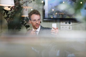 curly businessman in suit pointing with finger while taking selfie behind blurred window of cafe