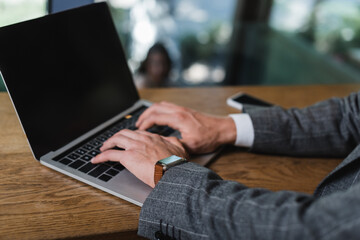 cropped view of businessman in suit typing on laptop with blank screen in cafe