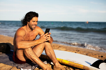 Portrait of handsome surfer with his surfboard. Young man using the phone while relaxing on the beach.