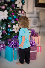 Fototapeta na wymiar A curly, blonde boy in a mint-colored T-shirt looks at the gifts under the Christmas tree.