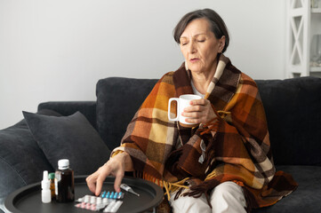 Senior woman has cold and flu, cover with blanket, sits on the couch and drinks hot remedy or tea, an older woman is treated at home, pills and medicines on the table near. Virus outspread concept