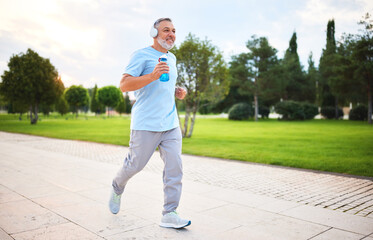 Happy senior man in headphones listening music while jogging outside in city park