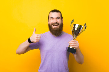 Cheerful male hipster is holding a trophy and showing like gesture over yellow background.