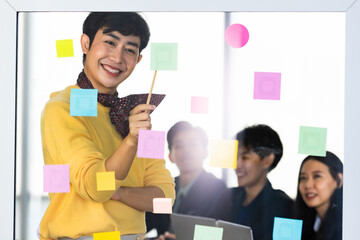 Asian trans woman on yellow shirt and scarf enjoy presenting business design to cheerful LGBT colleagues by point to colorful sticky paper note on glass board to explain creative strategy - obrazy, fototapety, plakaty